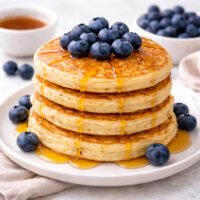 Four fluffy American blueberry pancakes stacked on a plate, golden and soft, fresh blueberries on the side, natural daylight, home-style breakfast.