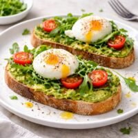 Two slices of avocado toast topped with poached eggs, cherry tomatoes and fresh arugula on a white plate, natural daylight brunch setting.