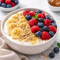 Yogurt berry bowl with banana slices, fresh berries, oats, chia seeds and honey in a ceramic bowl, natural daylight, no-cook breakfast.